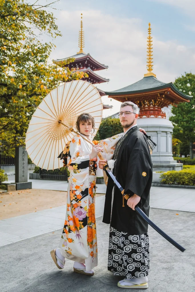 Couple in kimono rental attire at Asakusa Tokyo, woman in premium furisode with wagasa, man in montsuki hakama holding katana, five-story pagoda behind