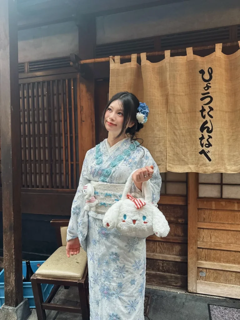 Girl in a white kimono with blue snowflake pattern holding a fluffy Cinnamoroll plush bag in front of a noren curtain in Asakusa, Tokyo