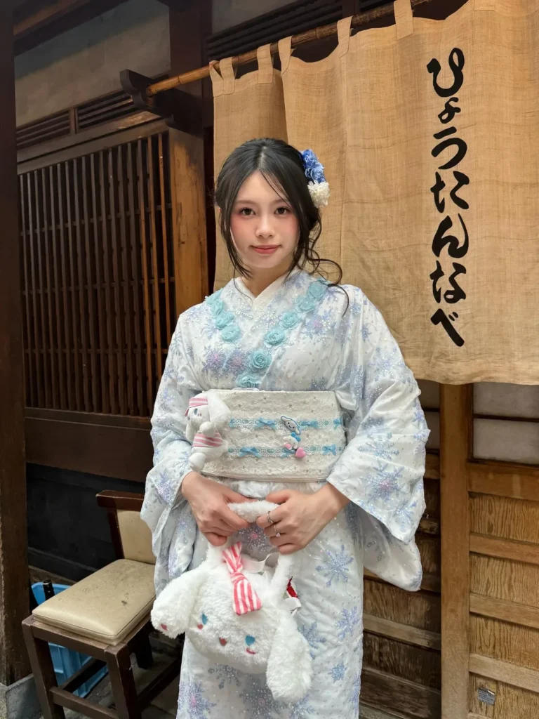 Girl in a white kimono with blue snowflake pattern holding a Cinnamoroll plush in front of a noren curtain in Tokyo