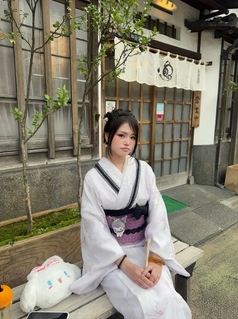 Girl in a white kimono with a purple Kuromi obi sitting on a bench beside a Cinnamoroll plush on an old street in Asakusa, Tokyo