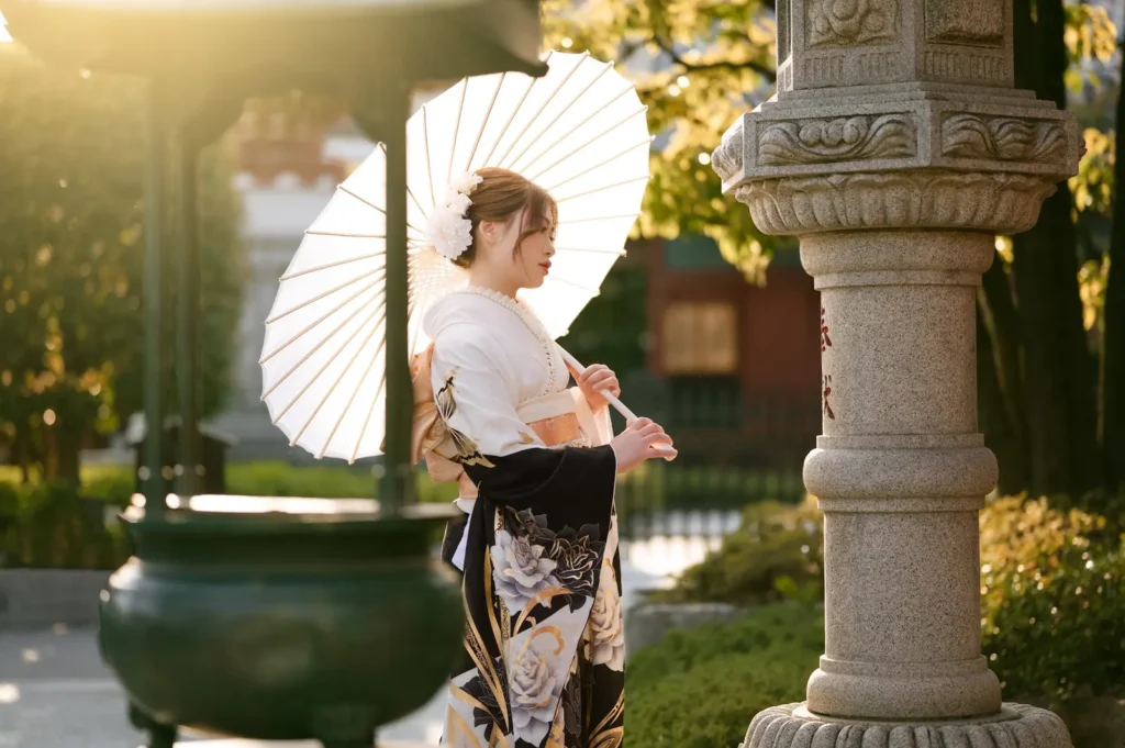 Guest in a white and black furisode holding a paper parasol beside a stone lantern at Sensoji in low golden light