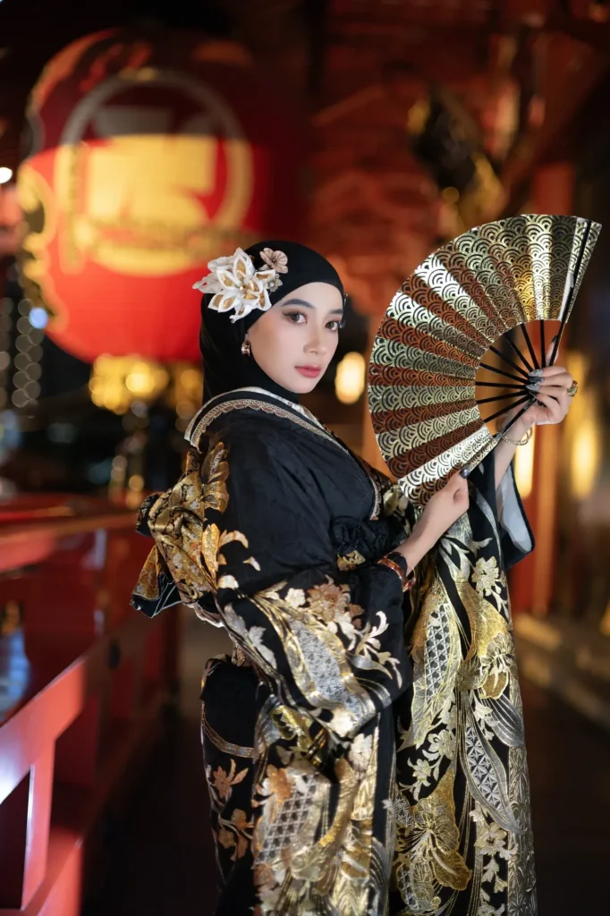 Guest in a black hijab and gold-patterned kimono holding a folding fan beside a glowing red temple lantern