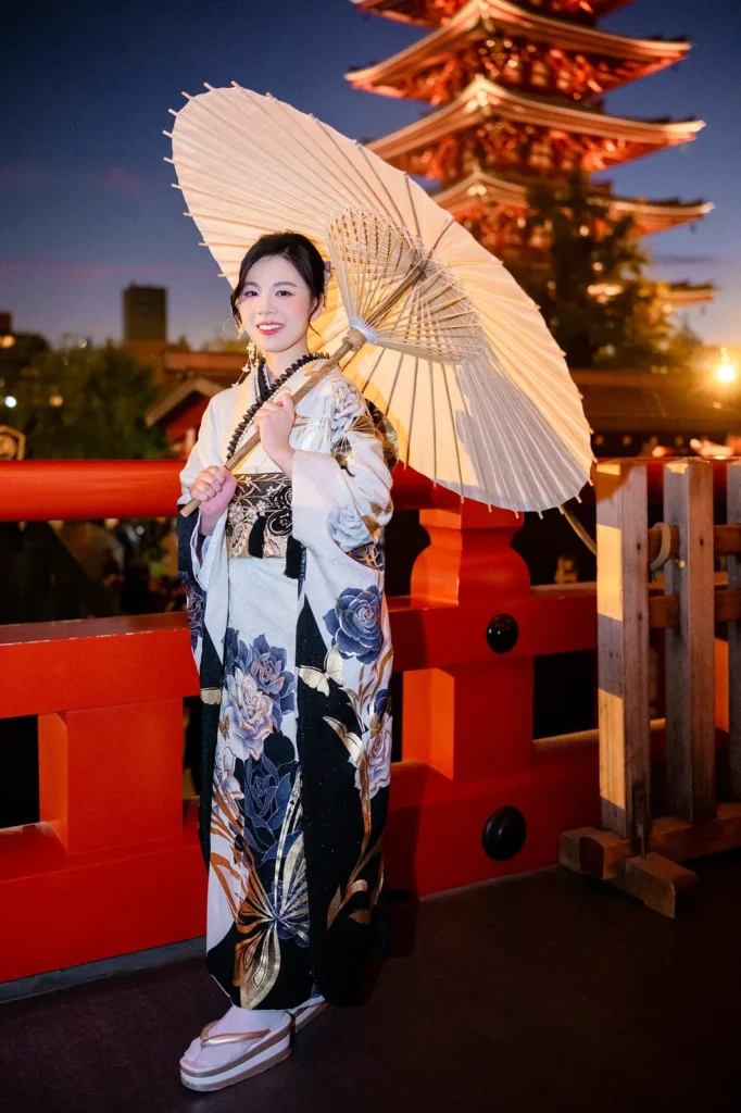 Woman in a black peony furisode holding a cream wagasa parasol on a red bridge at Sensoji Temple at dusk, with the five-story pagoda lit behind her