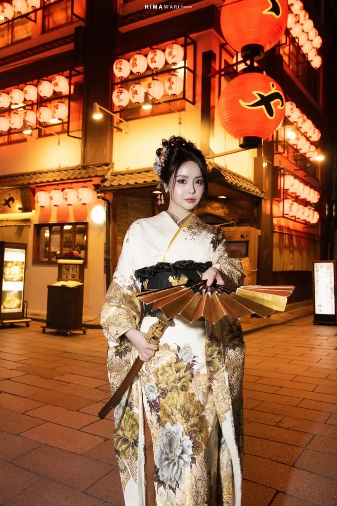 Woman in a cream and gold peony furisode holding a folding fan on a lantern-lit street in Asakusa at night
