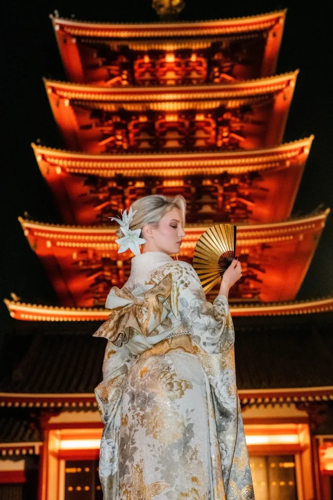 Woman in a premium gold and silver furisode holding a gold folding fan in front of the illuminated five-story pagoda at Sensoji Temple at night