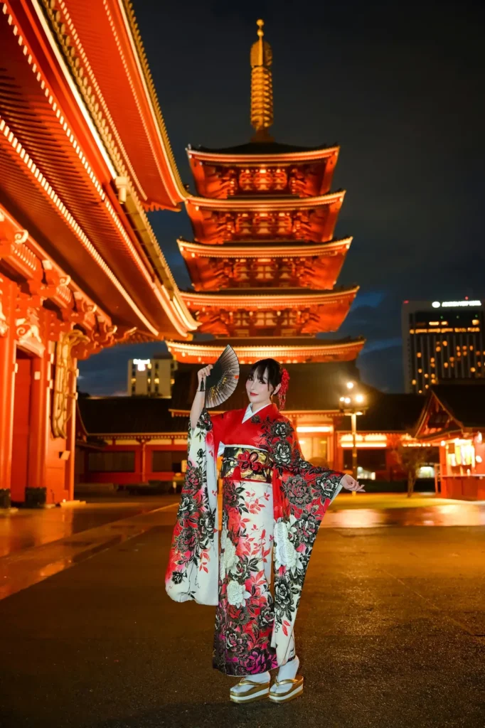 Woman in a red and black peony furisode holding a folding fan in front of the illuminated five-story pagoda at Sensoji Temple at night