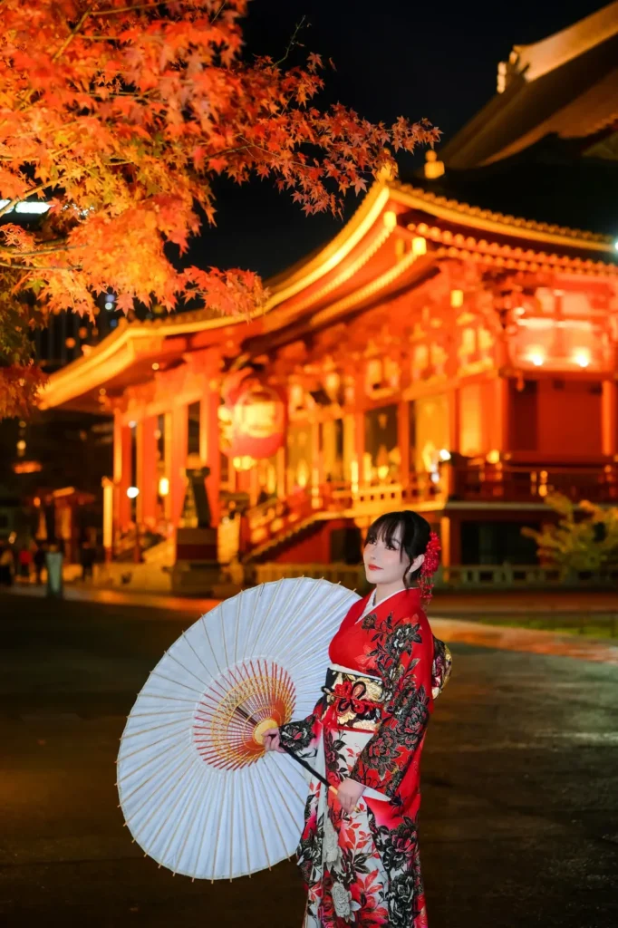 Woman in a red floral furisode holding a white wagasa parasol under autumn maple leaves in front of the illuminated Hozomon Gate at Sensoji, Asakusa