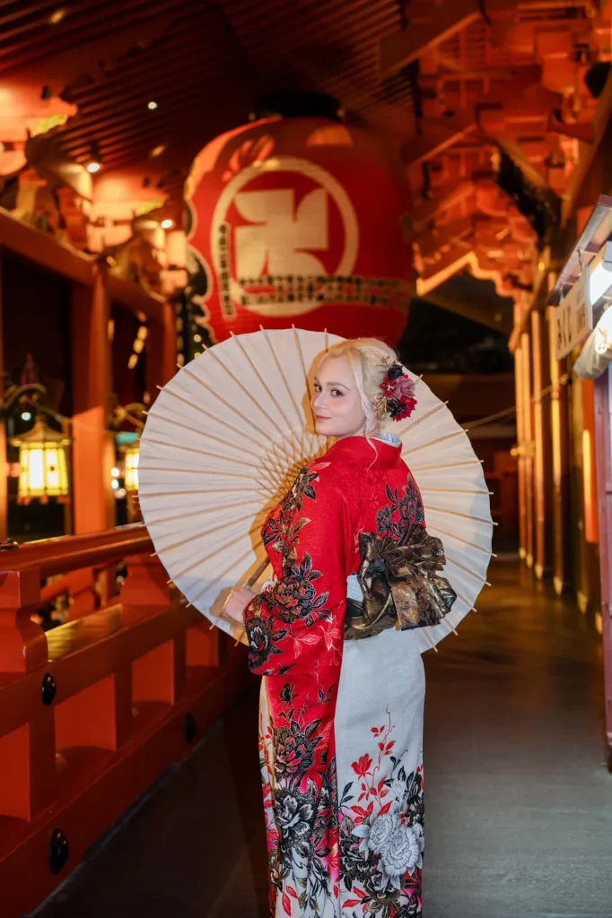 Woman in a red and white peony furisode holding a paper parasol in the lantern-lit corridor of Hozomon Gate at Sensoji Temple at night