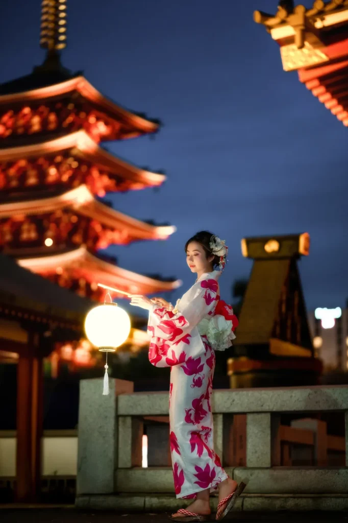 Woman in a white yukata with pink maple leaf pattern beside a glowing paper lantern at Sensoji Temple during blue hour in Asakusa