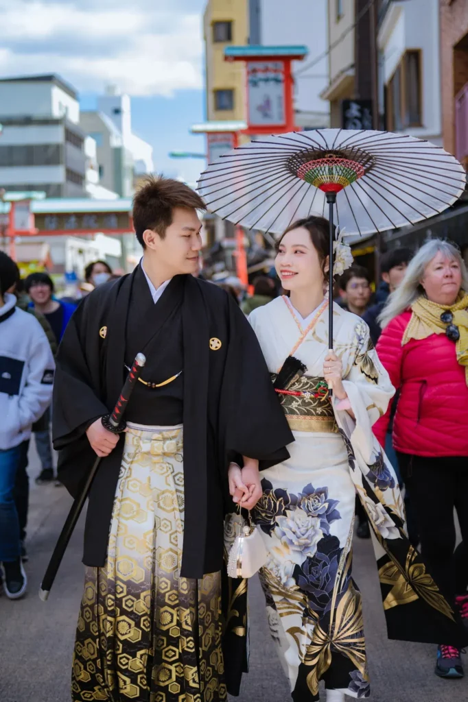 Couple in kimono walking a busy Asakusa shopping street, she holds a paper parasol, he wears a black haori