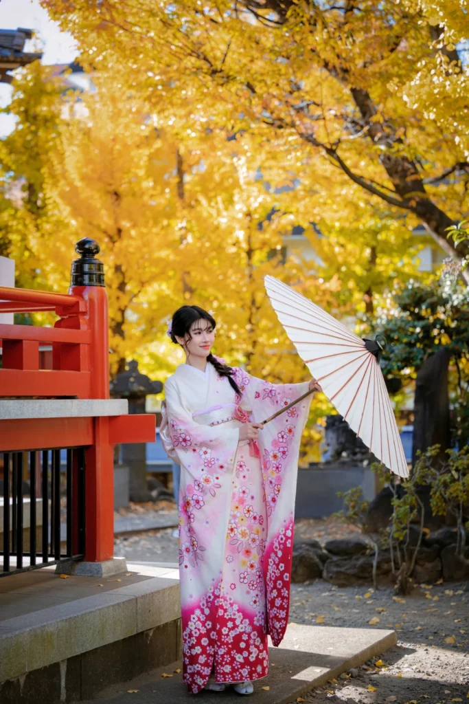 Woman in a pink and white furisode with a paper parasol under yellow ginkgo trees at Benten-dō, Asakusa