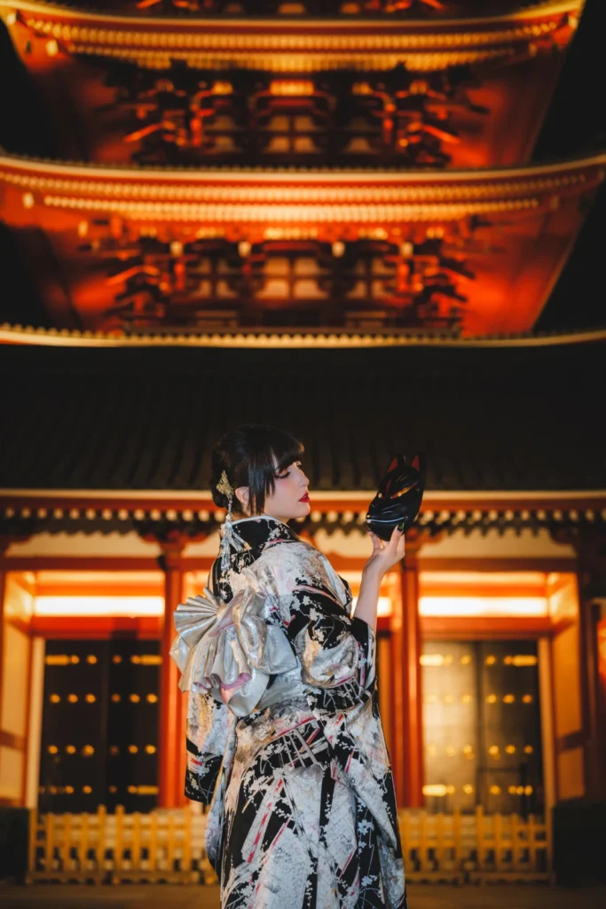 Woman in a black and white kimono looking up at the lit five-storied pagoda of Sensoji at night