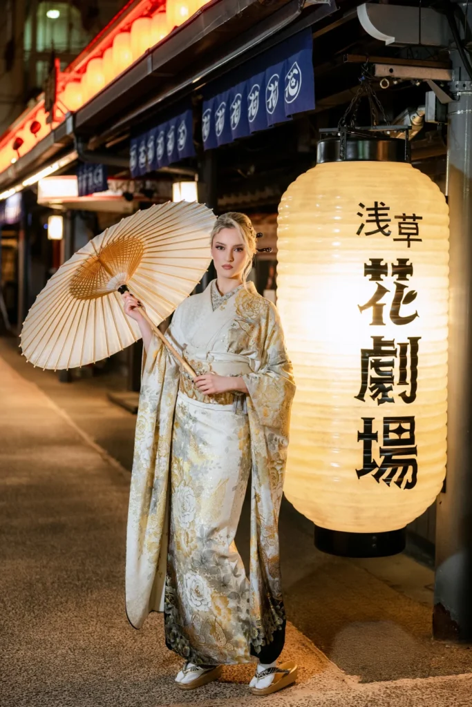 Woman in a cream kimono with a paper parasol beside a glowing theatre lantern on Hanayashiki Street, Asakusa