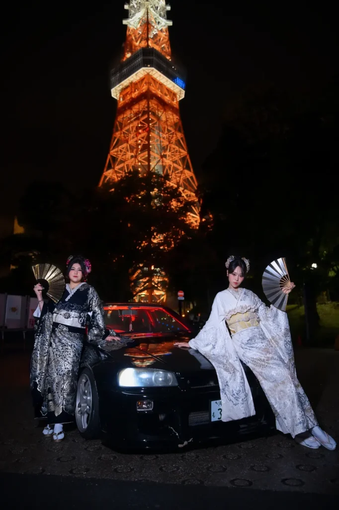 Two women in black-and-silver and white-and-gold furisode holding gold folding fans beside a black JDM sports car in front of the illuminated Tokyo Tower at night