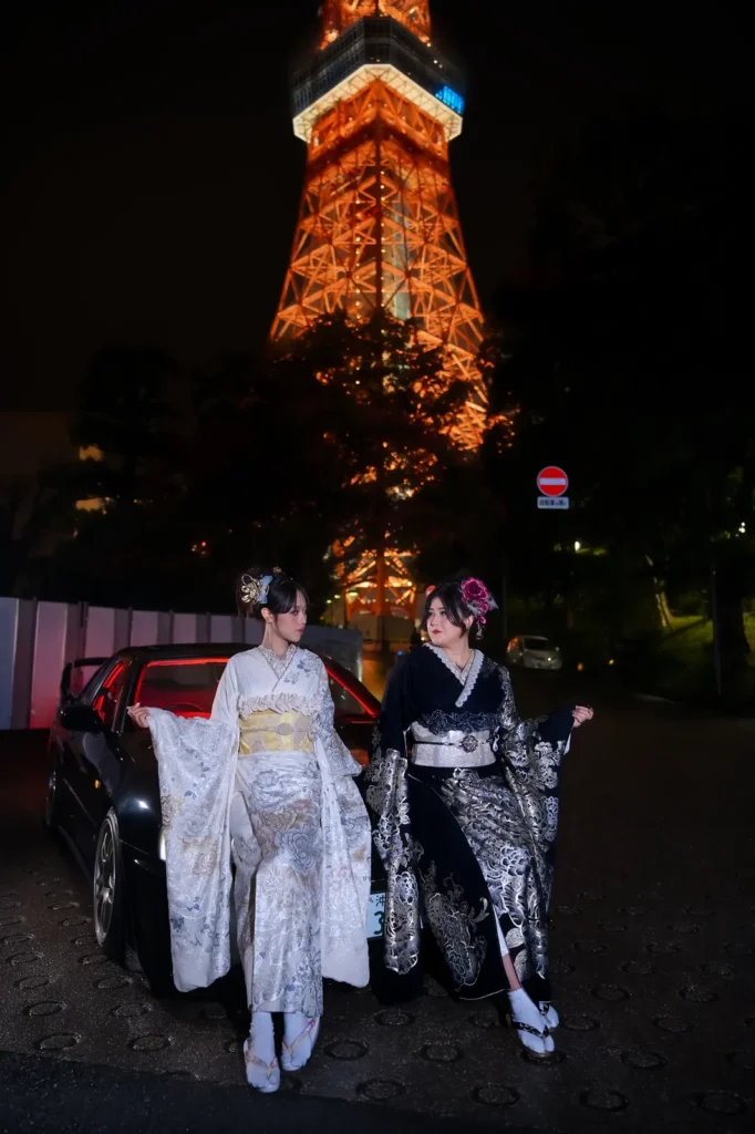 Two women in white and black kimono standing in front of a black JDM car with Tokyo Tower lit up behind them at night