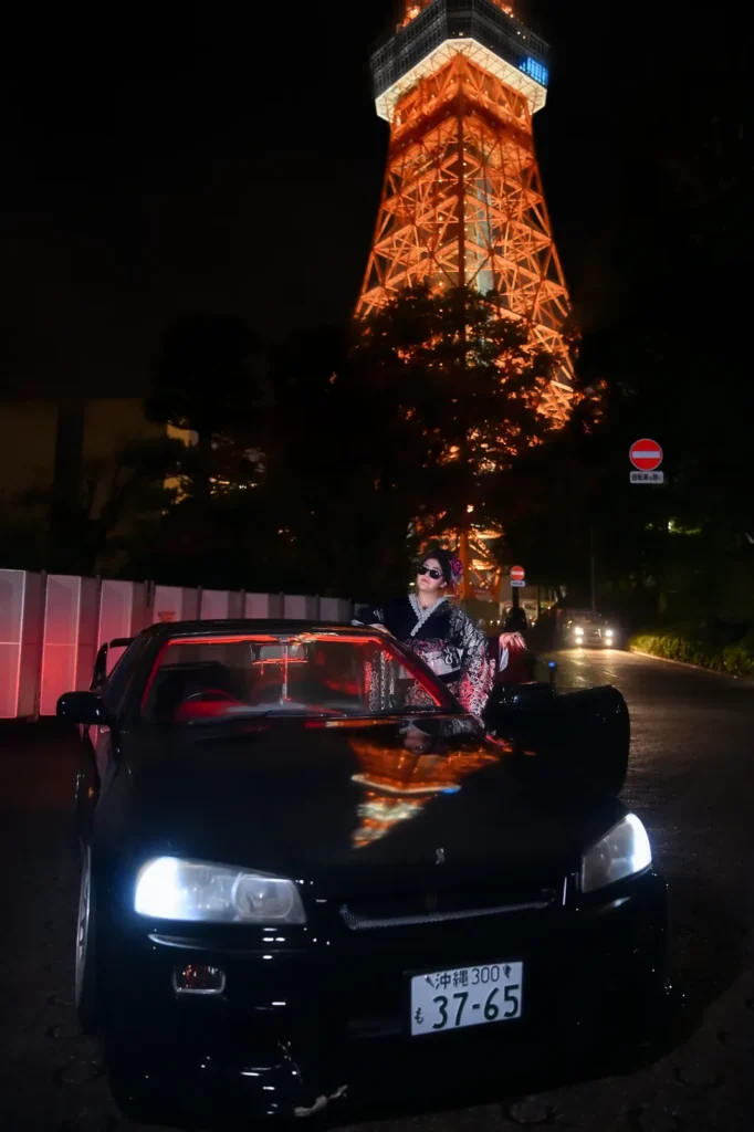 Woman in a black furisode on the bonnet of a black JDM car with headlights on below the illuminated Tokyo Tower