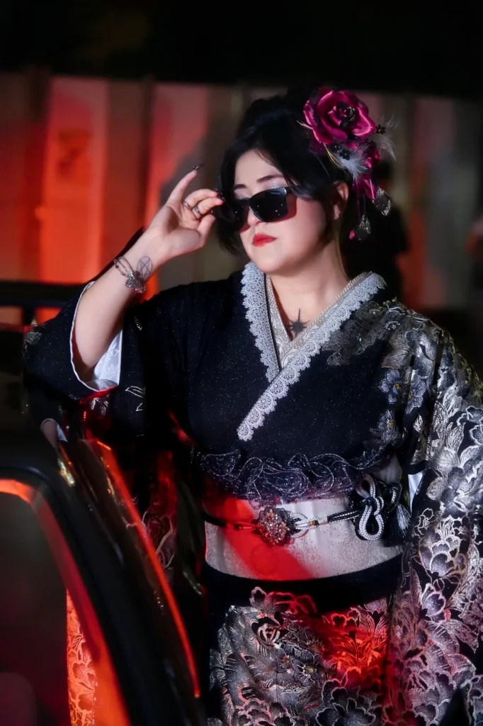 Woman in a black and silver lace kimono wearing sunglasses, leaning on the open door of a JDM car in red light
