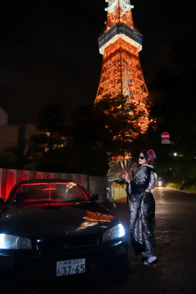 Woman in a black and silver furisode holding a gold fan beside a black JDM car at night, Tokyo Tower lit behind her