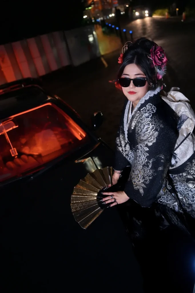 Woman in a black and silver kimono with sunglasses and a gold folding fan posing by a car in red tail-light glow