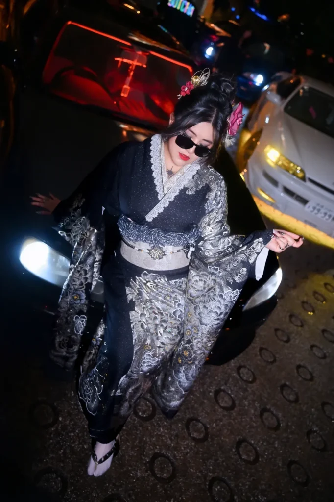 Low-angle shot of a woman in a black and silver kimono wearing sunglasses beside a JDM car with headlights on