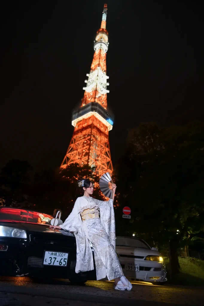 Woman in a white and gold furisode posing between two JDM cars at the foot of the illuminated Tokyo Tower