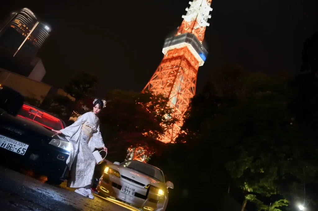 Wide shot of a woman in a white furisode beside JDM cars with Tokyo Tower glowing orange at night