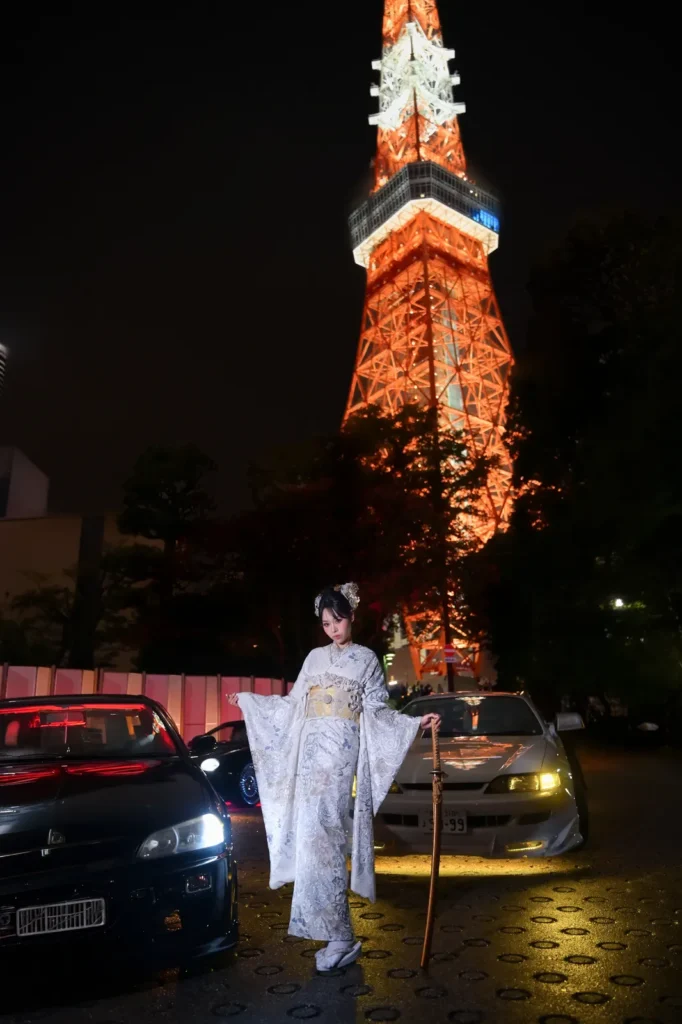 Woman in a white and gold furisode standing beside JDM cars under the illuminated Tokyo Tower at night