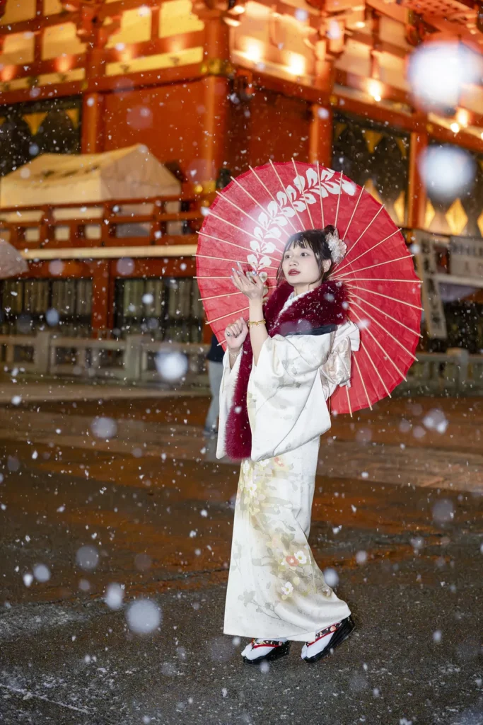 Woman in a cream kimono and red winter shawl holding a red parasol as snow falls at Sensoji, Asakusa